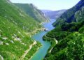 A scenic road winding through a lush green valley, with the turquoise Neretva River flowing alongside and majestic mountains in the background.