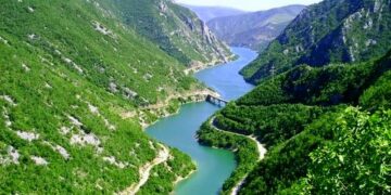 A scenic road winding through a lush green valley, with the turquoise Neretva River flowing alongside and majestic mountains in the background.