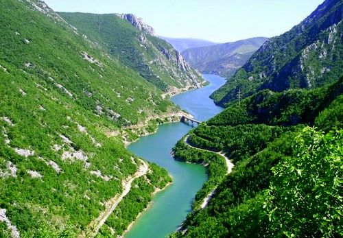 A scenic road winding through a lush green valley, with the turquoise Neretva River flowing alongside and majestic mountains in the background.