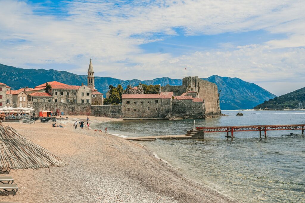 Budva, Montenegro — Aged church and building facades near sandy shore and ocean with dock behind ridge under cloudy sky
