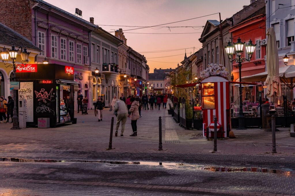 Novi Sad, Serbia — Captivating street scene in Novi Sad, Serbia, during twilight with illuminated shops and lively foot traffic.