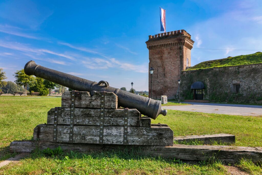 Osijek, Croatia — A vintage cannon rests before a historic tower in Osijek, Croatia under a clear blue sky.